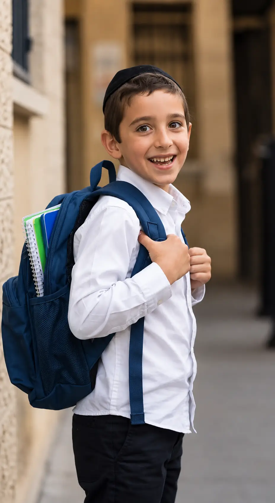 A smiling boy in a kippah and white shirt on his way to school, a navy backpack on his shoulders
